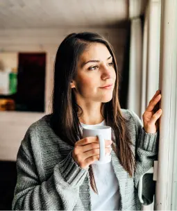 Young woman in a gray cardigan holding a white mug, gazing thoughtfully out the window with a calm, reflective expression in a cozy home setting.