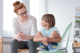 Female therapist in glasses talking with a young boy seated on a green chair, both engaged in a warm conversation during a counseling session.