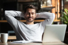 Relaxed young man with hands behind his head sitting at a table with a laptop, smiling and looking content during a break in a cozy cafe setting.