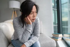 Worried middle-aged woman sitting on a sofa, resting her chin on her hand and looking out the window with a sad, thoughtful expression.